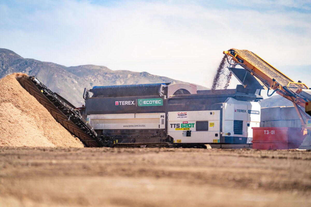 A Terex Ecotec TTS 620T trommel screen sorts and processes mulch material at an outdoor site, with a conveyor discharging fine material onto a pile. Mountain landscape in the background under a bright sky.