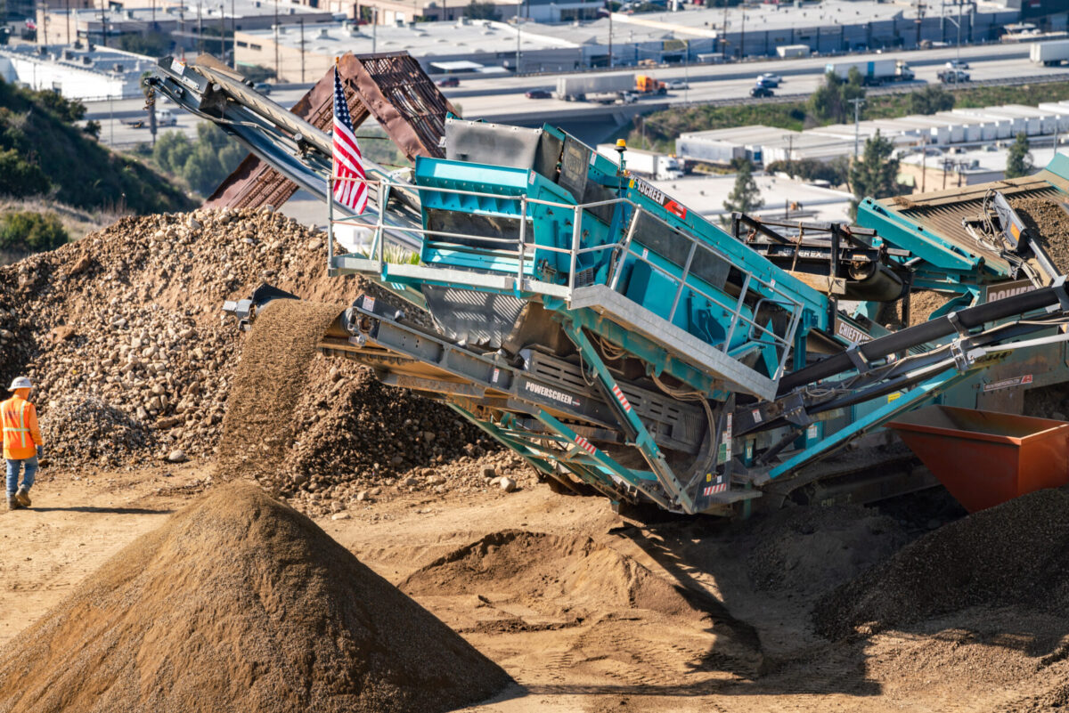 A Powerscreen Chieftain screener sorts aggregates on a construction site, with material flowing from the conveyor into a pile. A worker in an orange safety vest and hard hat observes the process, while an American flag is displayed on the machine.