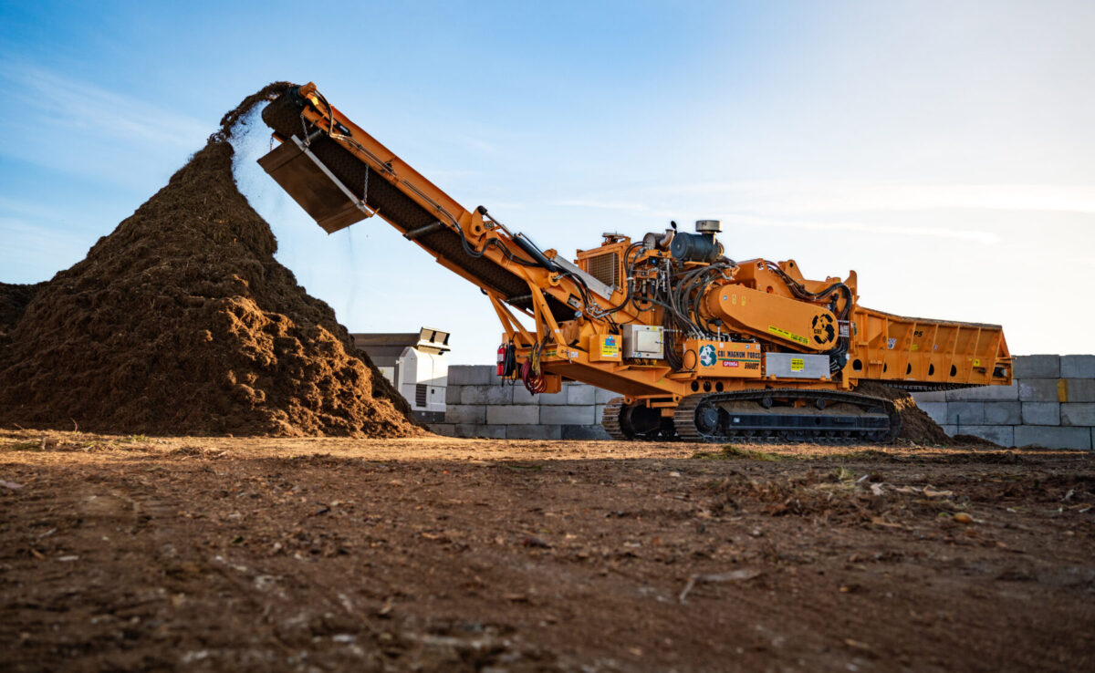 A large industrial horizontal grinder processes wood waste, discharging mulch material onto a tall pile. The machine, painted orange with various warning labels and company logos, operates on crawler tracks in an outdoor yard with concrete block walls in the background under a clear blue sky.