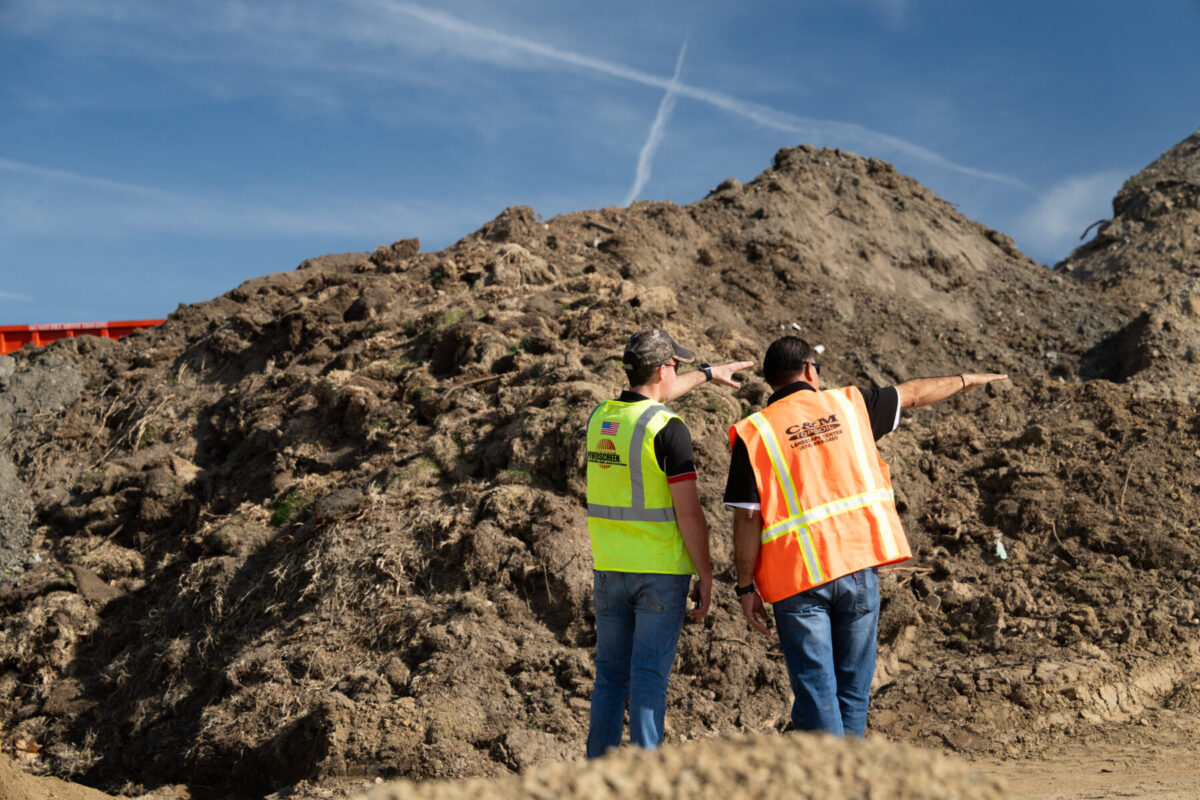 Two workers in safety vests stand in front of a large mound of soil and debris, pointing toward the material as they discuss the job site. Blue sky with contrails visible above.