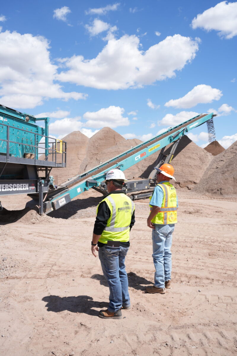 Two people in safety ppe standing in front of a Powerscreen crusher and conveyor working