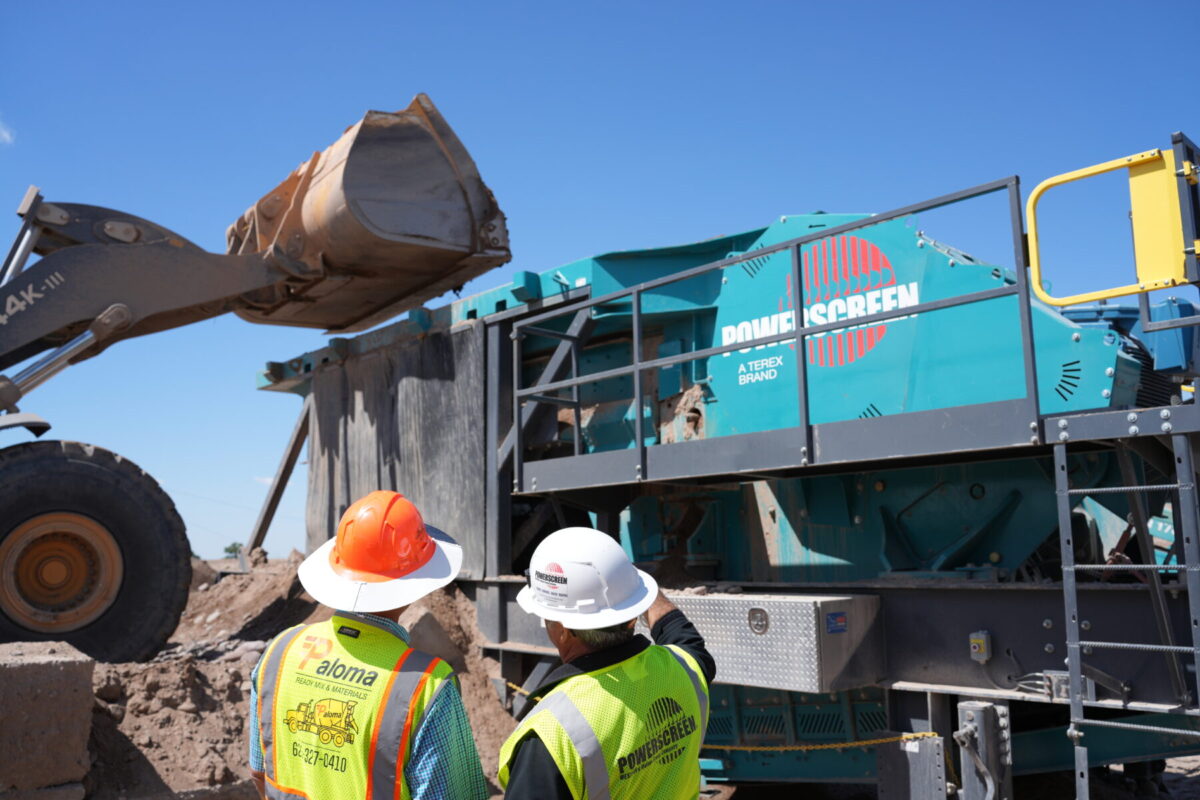 Two people in safety ppe standing in front of a Powerscreen Gladiator PT400 jaw crusher while a wheel loader is loading the hopper