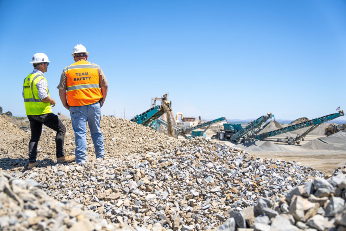 Dos hombres con chalecos de seguridad y cascos mientras están de pie sobre una pila de áridos con vistas a una operación de trituración y cribado de Powerscreen.