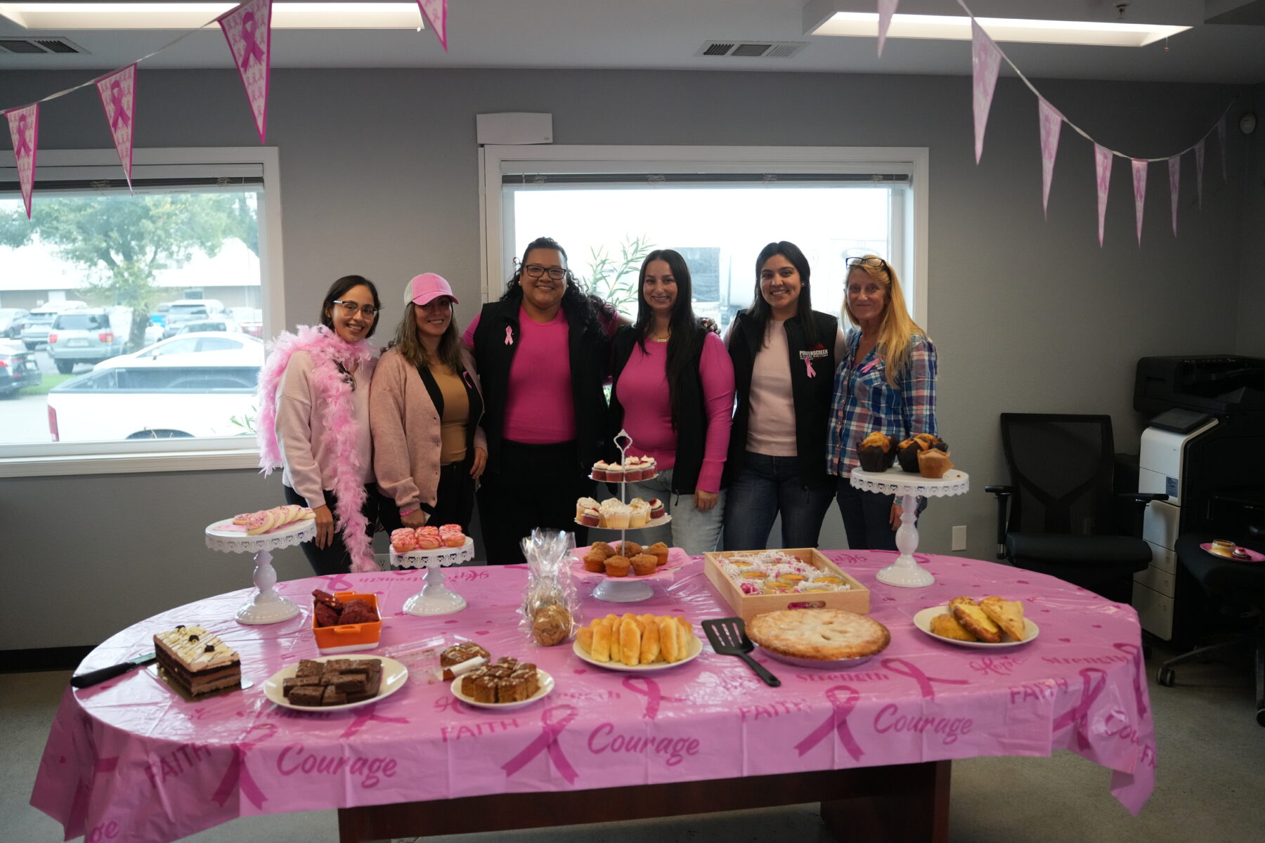 Six women in pink standing in front of a table of baked goods for a bake sale for breast cancer awareness month.